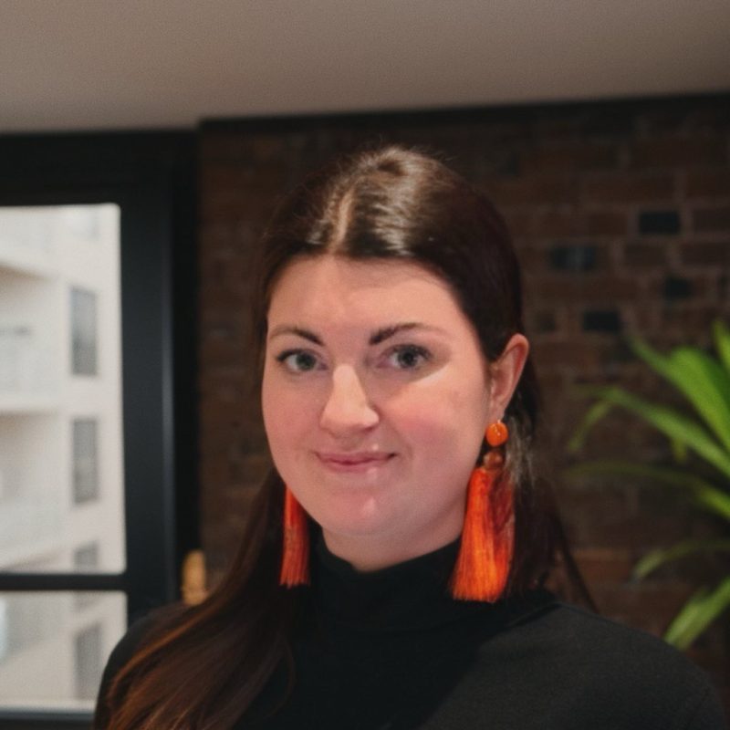 A woman with long brown hair wears a black t-shirt and vibrant orange earrings.