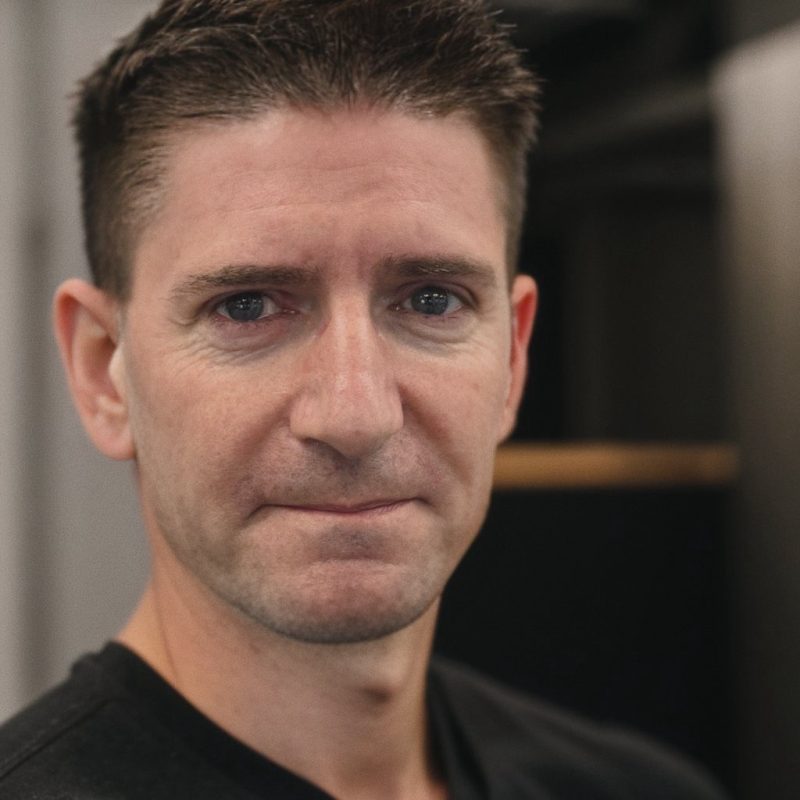 A man with short brown hair and blue eyes wears a black V-neck T-shirt and has a slight smile.