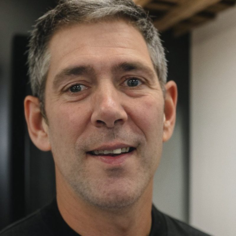 A man with short silver and black hair and blue eyes wears a black sweatshirt and has a happy smile.