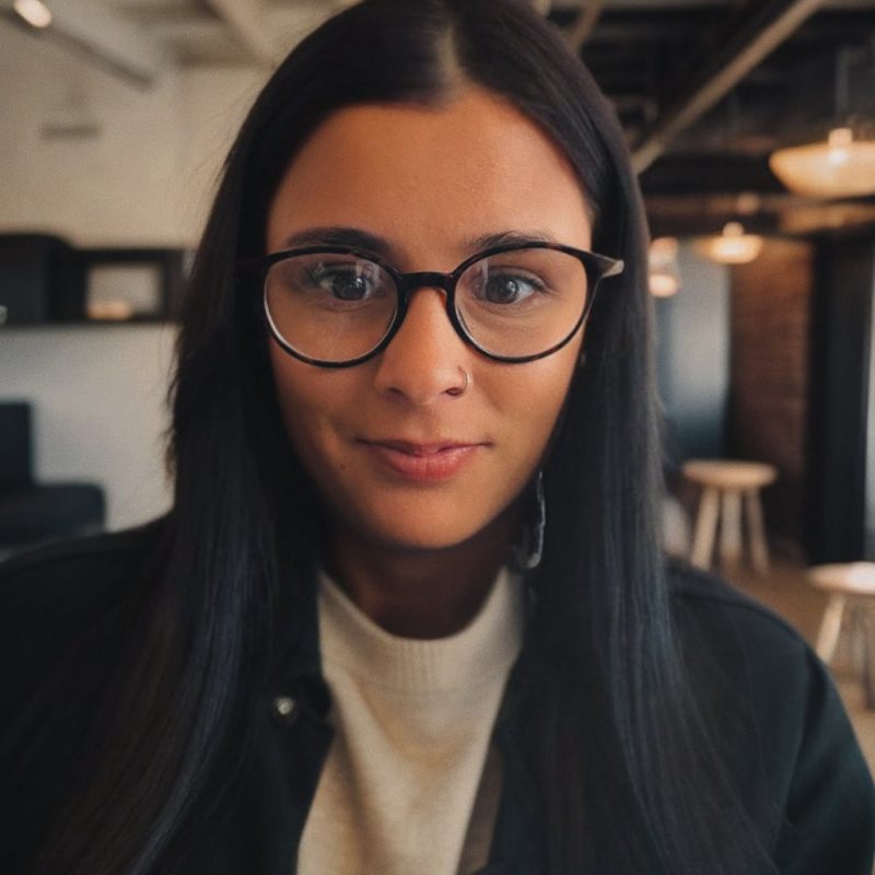 A lady with dark hair, glasses, a white jumper, a black jacket, and a nose ring.