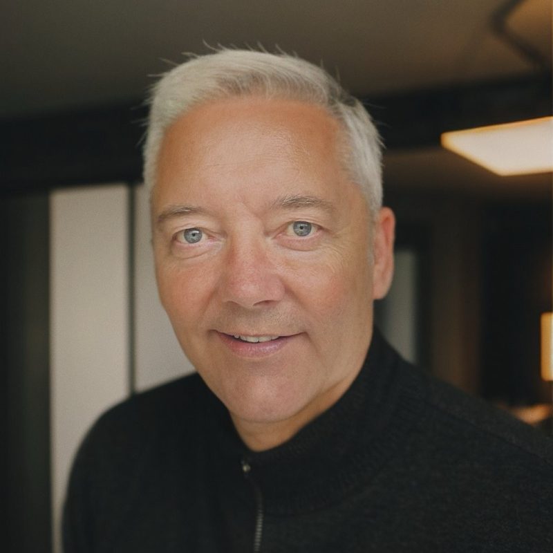 A man with short silver hair and a black zipped jacket is smiling, with light blue-grey eyes.